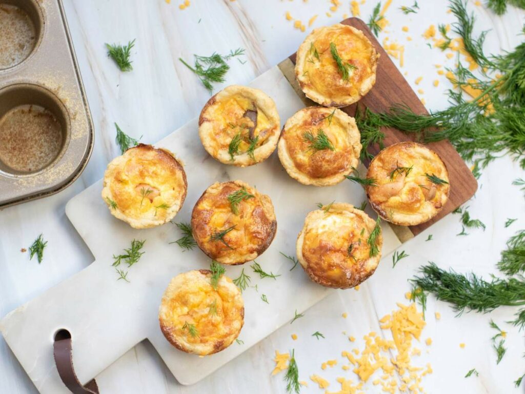 Seven bsmoked salmon tartlets topped with herbs are arranged on a white cutting board.