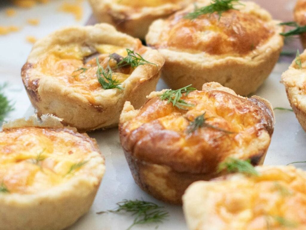 Several smoked salmon tartlets with golden, flaky crusts are topped with fresh dill and arranged on a light surface.
