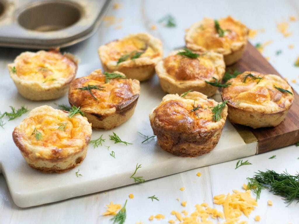 Eight smoked salmon tartlets topped with fresh dill are arranged on a marble board, with scattered shredded cheese and herbs around them.