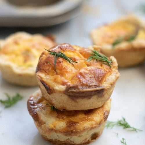Two smoked salmon tartlets are stacked on a white surface, garnished with fresh dill.