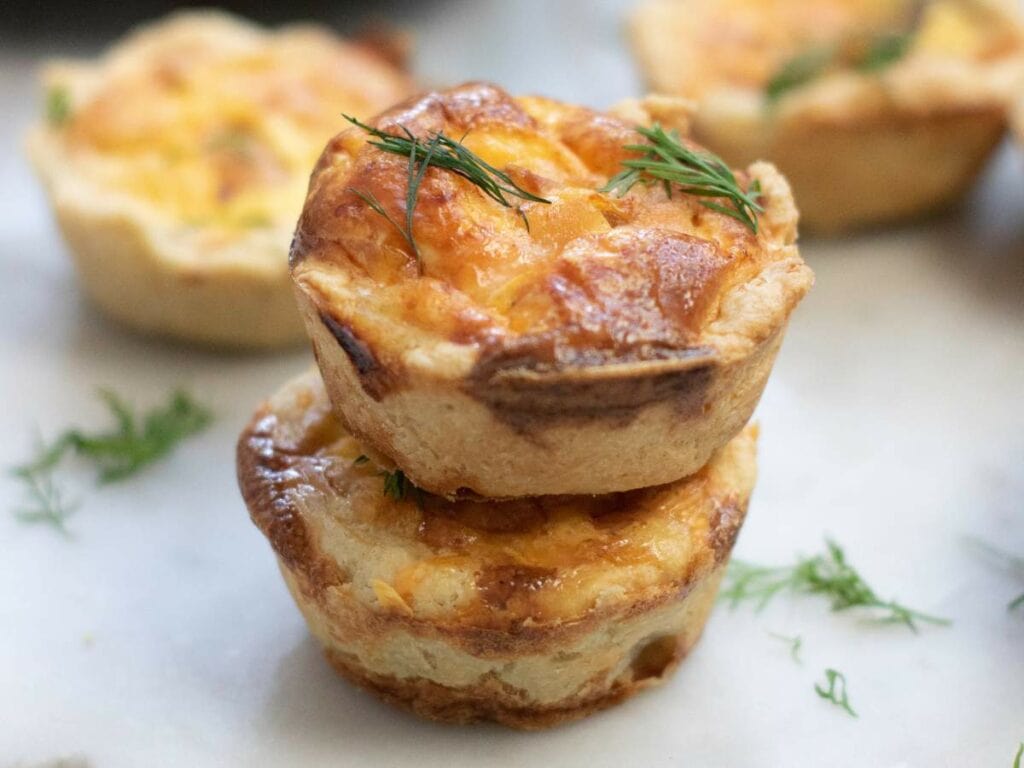 Two smoked salmon tartlets are stacked on a marble surface, garnished with small sprigs of fresh dill.