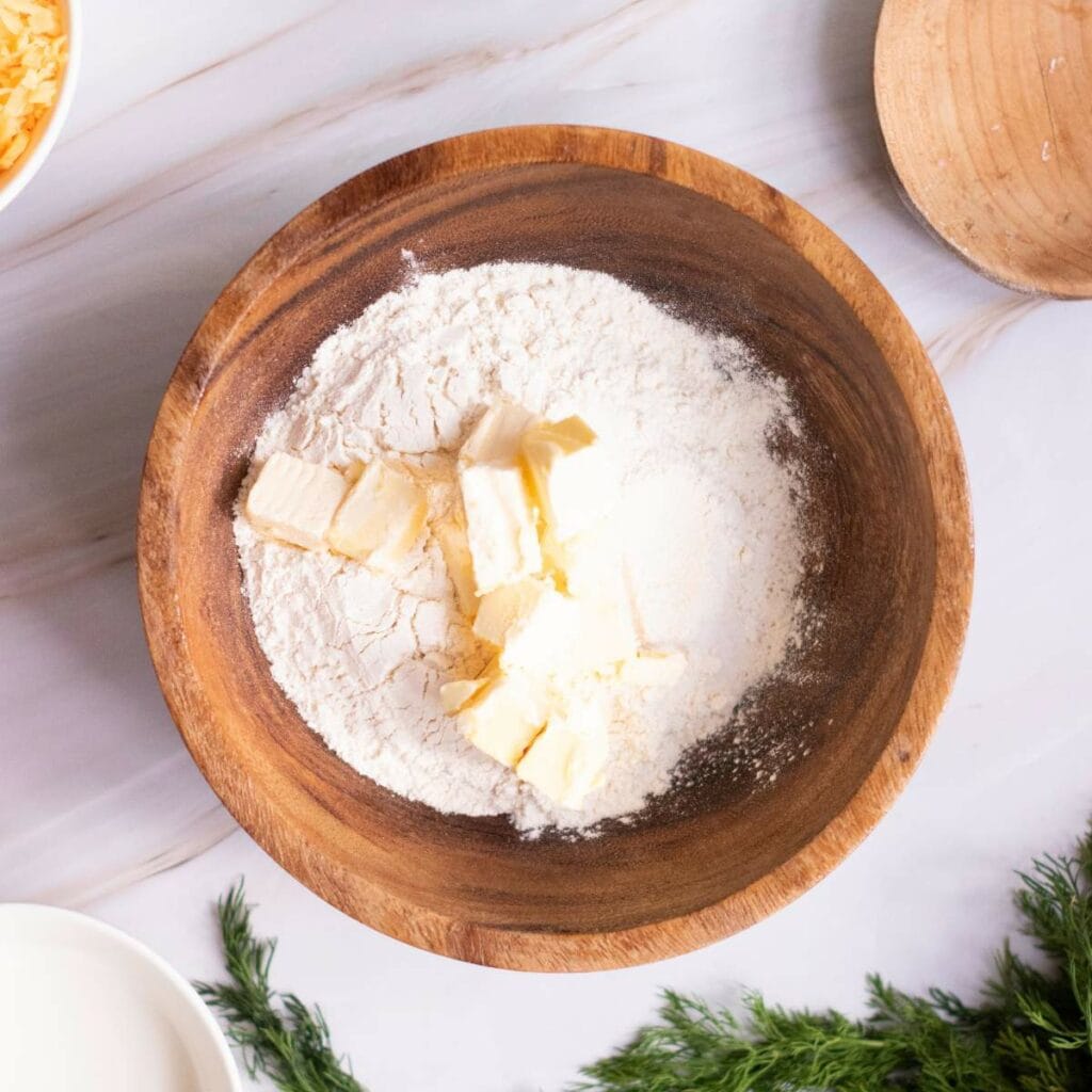 A wooden bowl containing flour and chunks of butter sits on a marble surface.