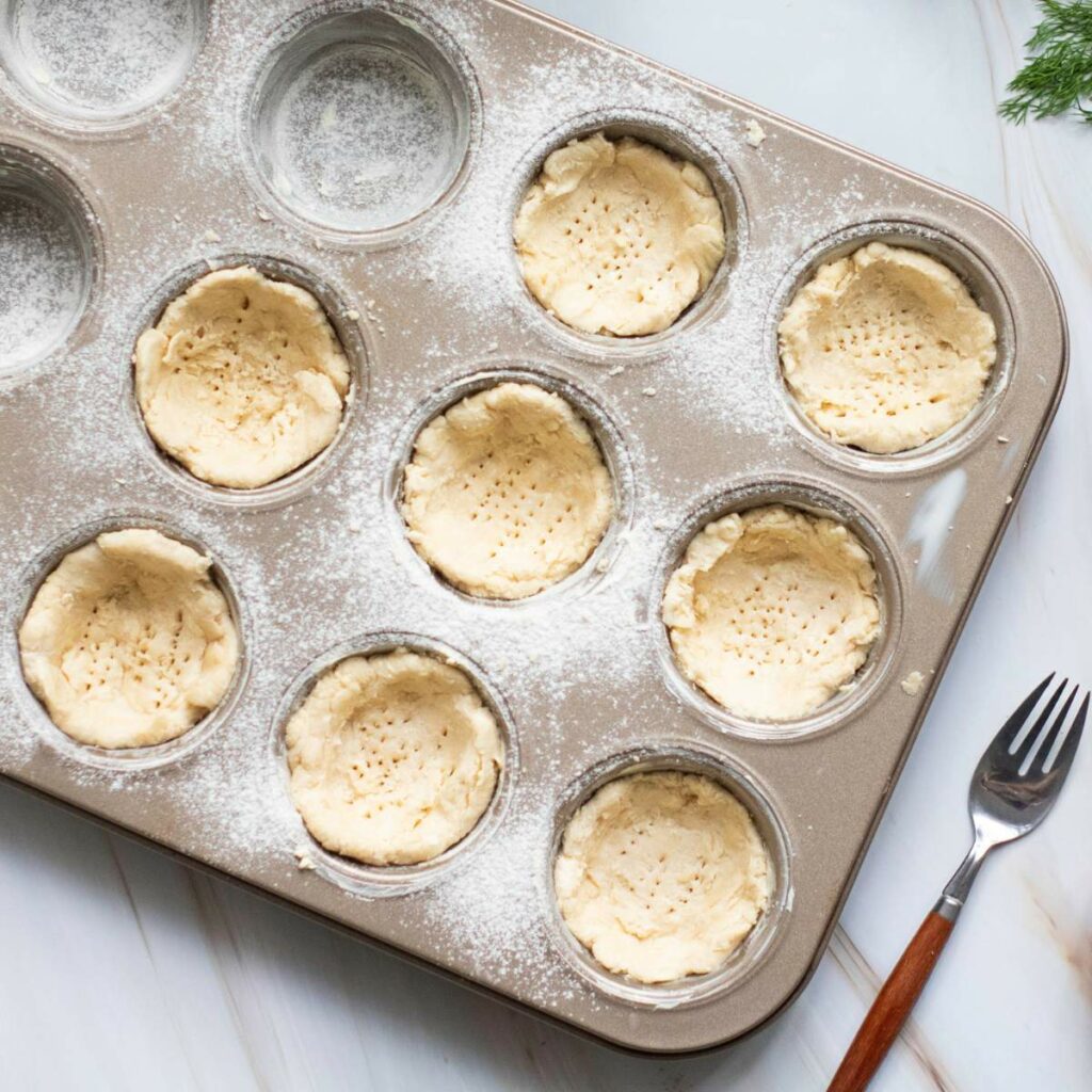 Muffin tin with nine unbaked pie crusts, each docked with a fork, sits on a floured surface.