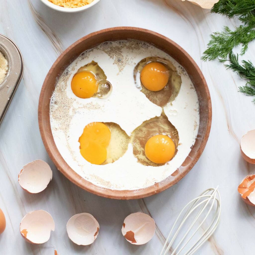 A ceramic bowl with cracked eggs and cream, surrounded by eggshells, a whisk, herbs, and a muffin tin.