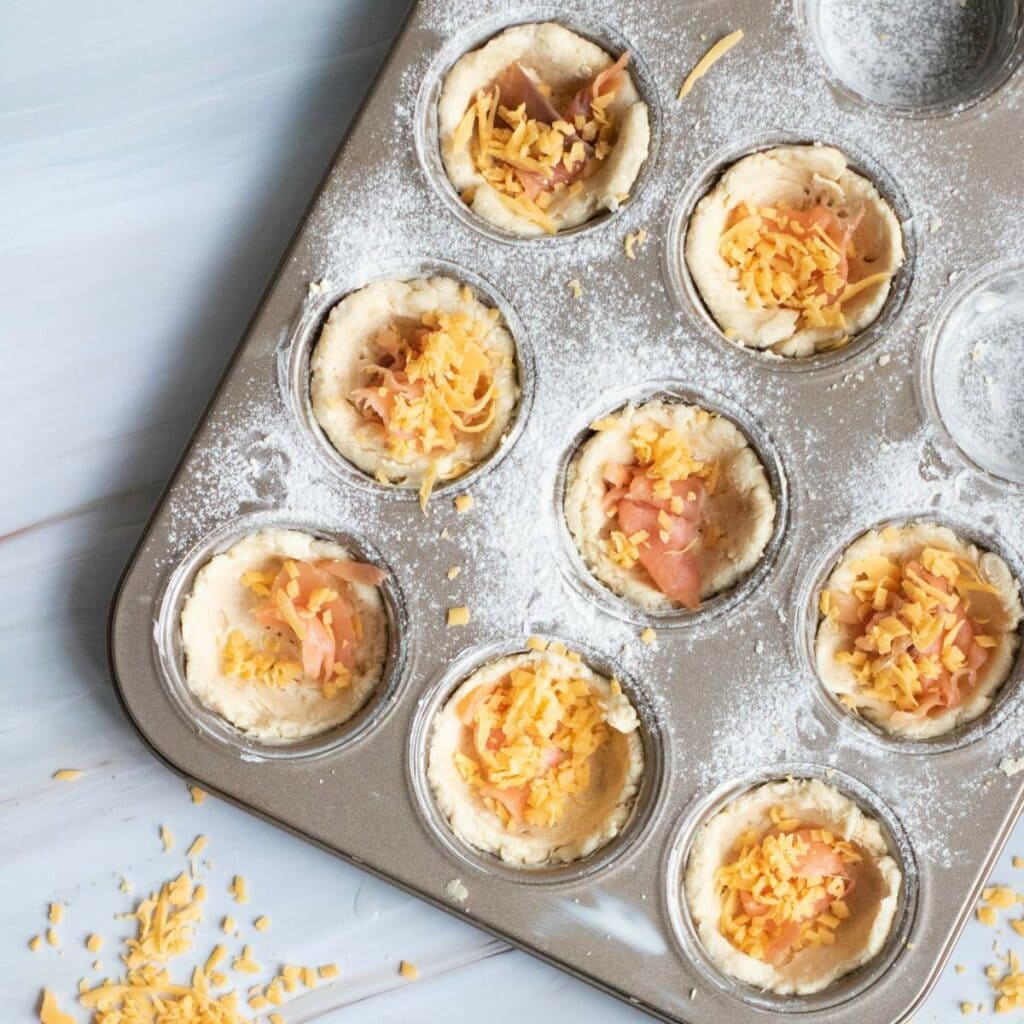 A muffin tin filled with dough cups, some containing grated cheese and sliced ham.