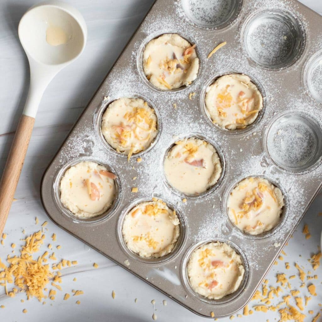 A muffin tin filled with unbaked batter topped with shredded cheese, some batter splatters, and a spoon.