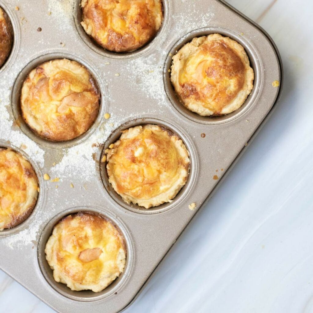 A close-up of a muffin tin filled with six baked egg tarts placed on a white marble surface.