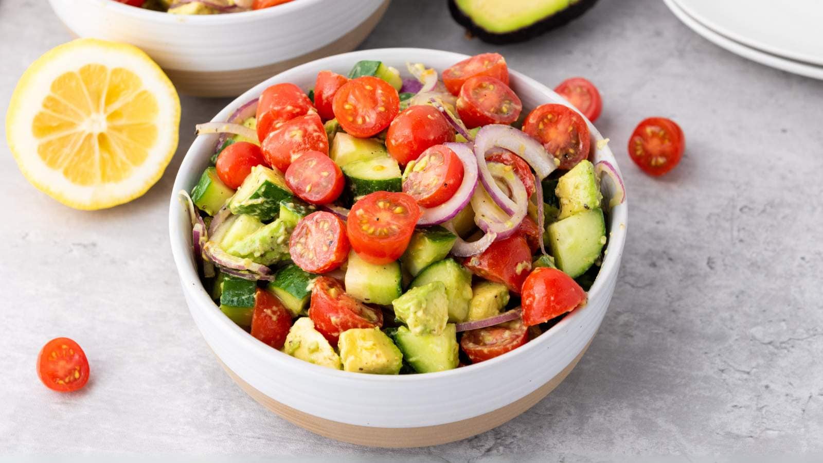 A bowl of summer salad with cherry tomatoes, avocado, cucumber, red onion, and lemon on a light surface.
