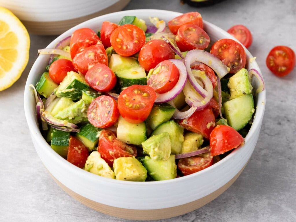 A bowl of summer salad with cherry tomatoes, cucumber, avocado, and red onion slices on a light gray surface.