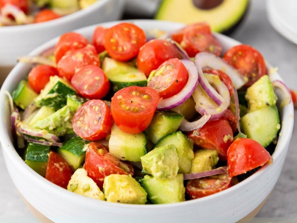 A bowl of summer salad containing cherry tomatoes, cucumber, avocado, and red onion slices.