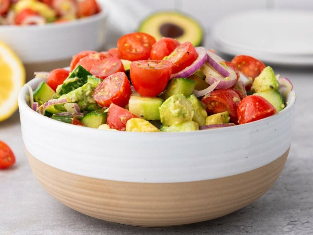 A bowl of summer salad containing cherry tomatoes, cucumber, avocado, and red onion slices on a light-colored surface.