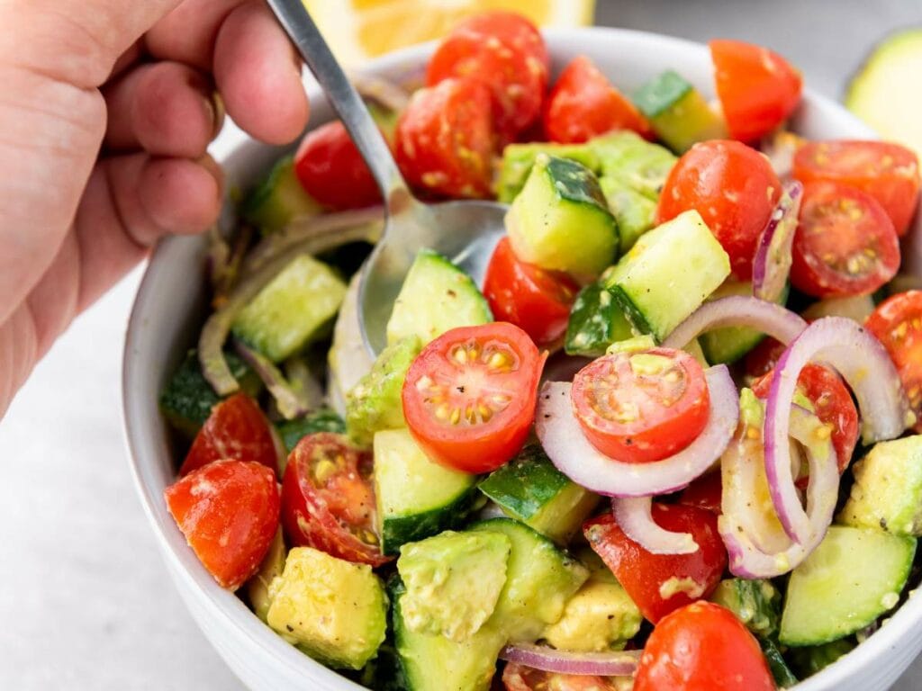 A hand holds a spoon in a bowl of summer salad containing cherry tomatoes, cucumber, avocado, and red onion slices.