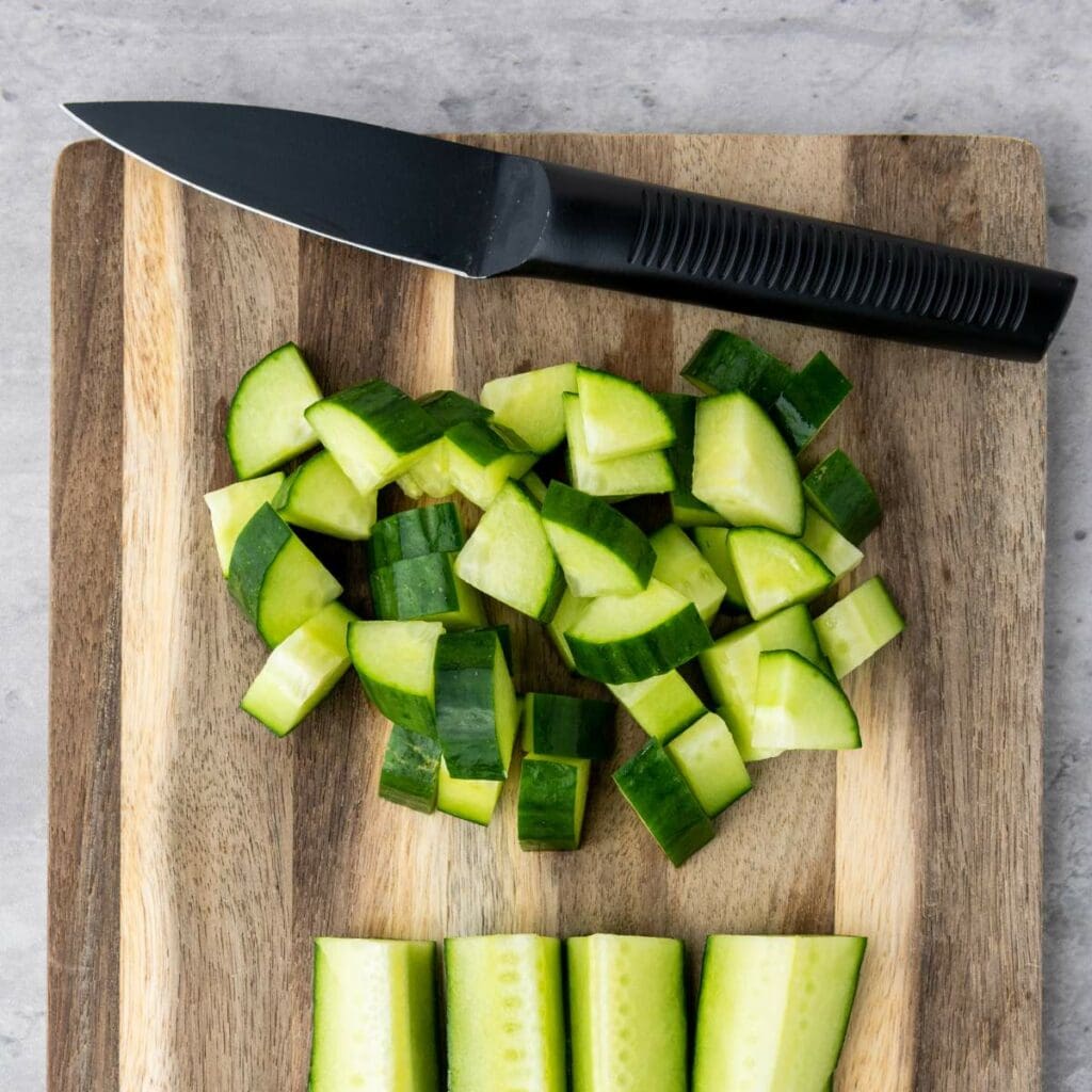 A black-handled knife and chopped cucumber pieces on a wooden cutting board.