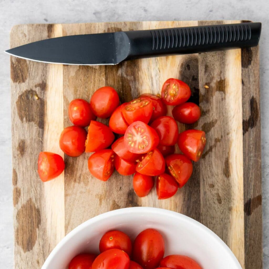 A pile of halved cherry tomatoes and a black-handled knife are on a wooden cutting board, with a white bowl of whole tomatoes in the foreground.