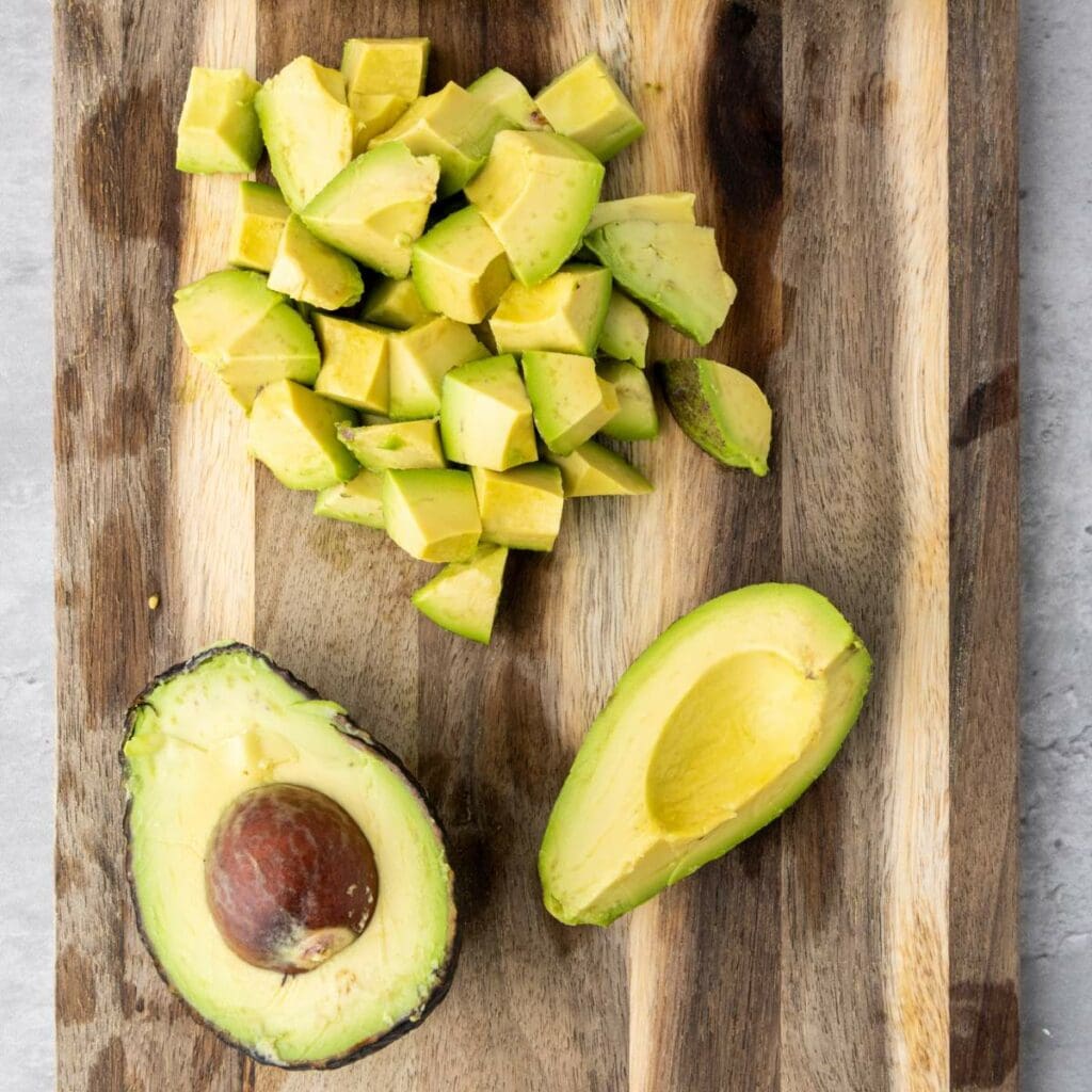 Diced avocado pieces and two avocado halves, one with the pit, on a wooden cutting board.