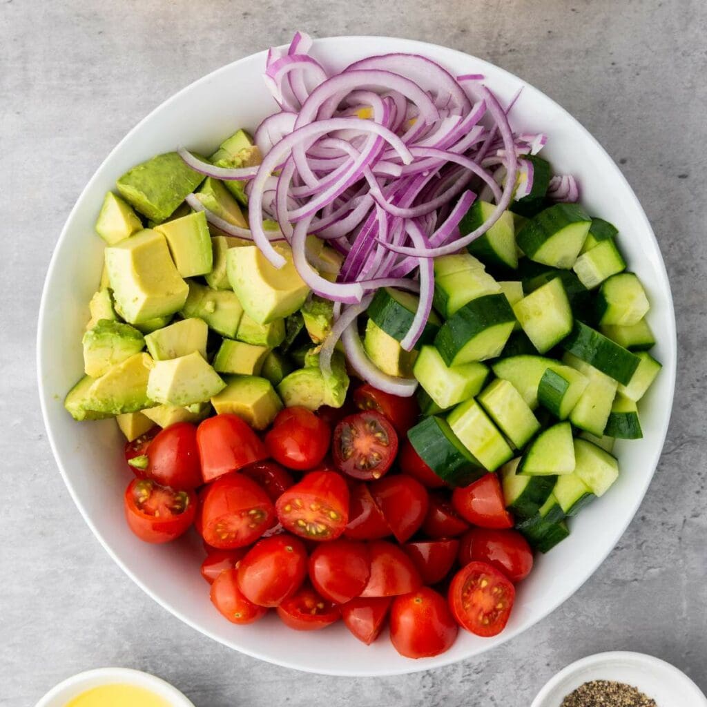 A white bowl containing chopped avocado, sliced red onion, diced cucumber, and halved cherry tomatoes, arranged in separate sections.
