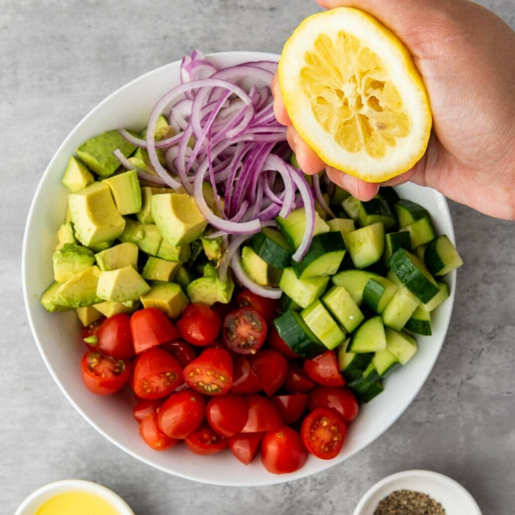 A hand squeezes a lemon over a bowl containing sliced red onion, avocado, cucumber, and cherry tomatoes.
