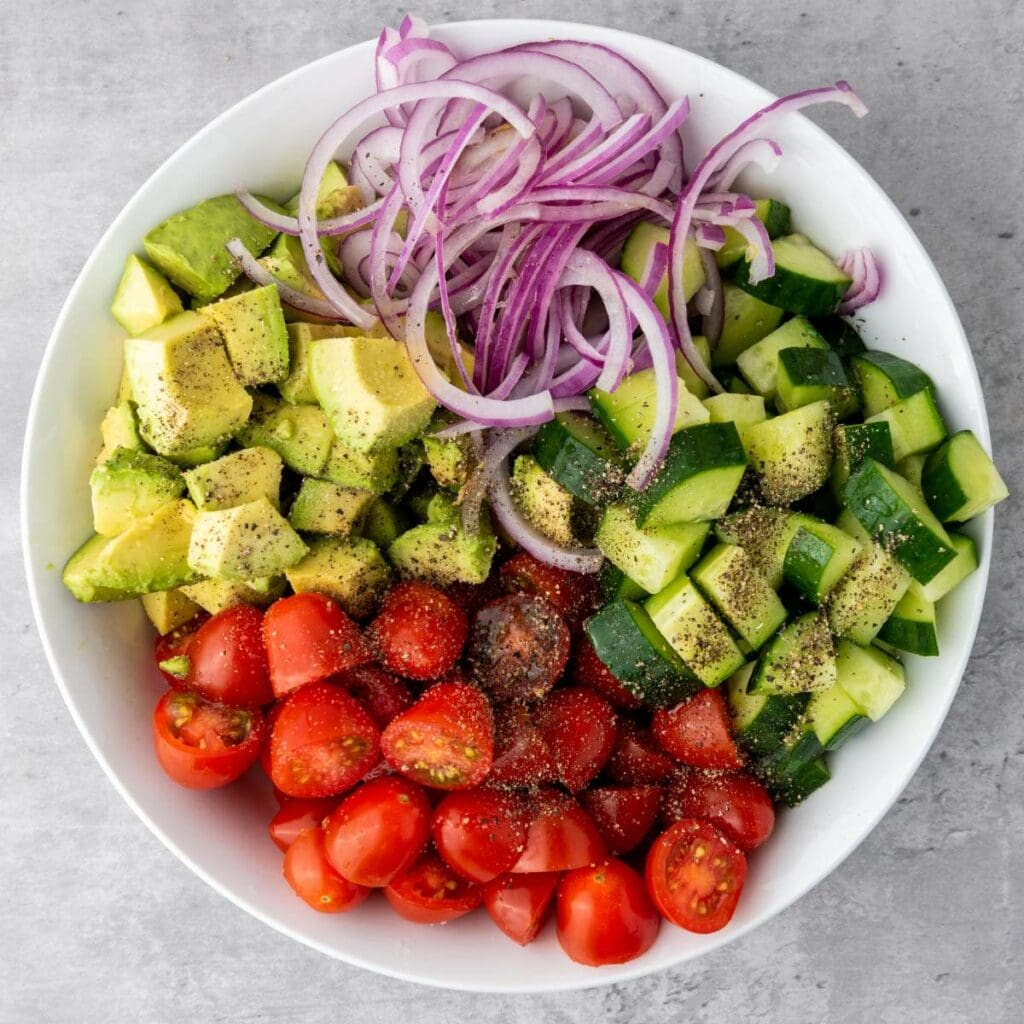 A white bowl filled with chopped avocado, red onion, cucumber, and cherry tomatoes.