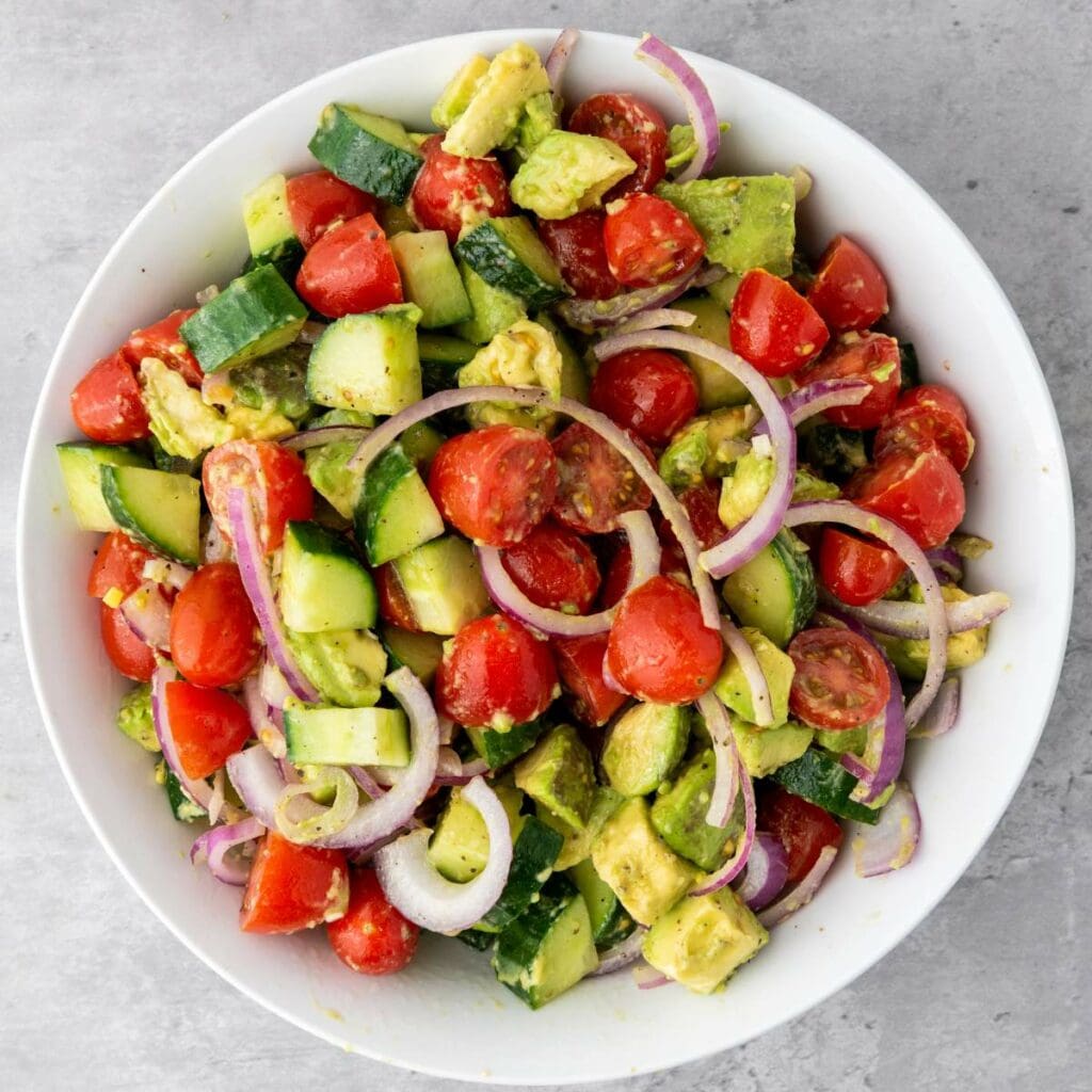 A white bowl filled with a salad of cherry tomatoes, cucumber, avocado, and sliced red onion on a light gray surface.