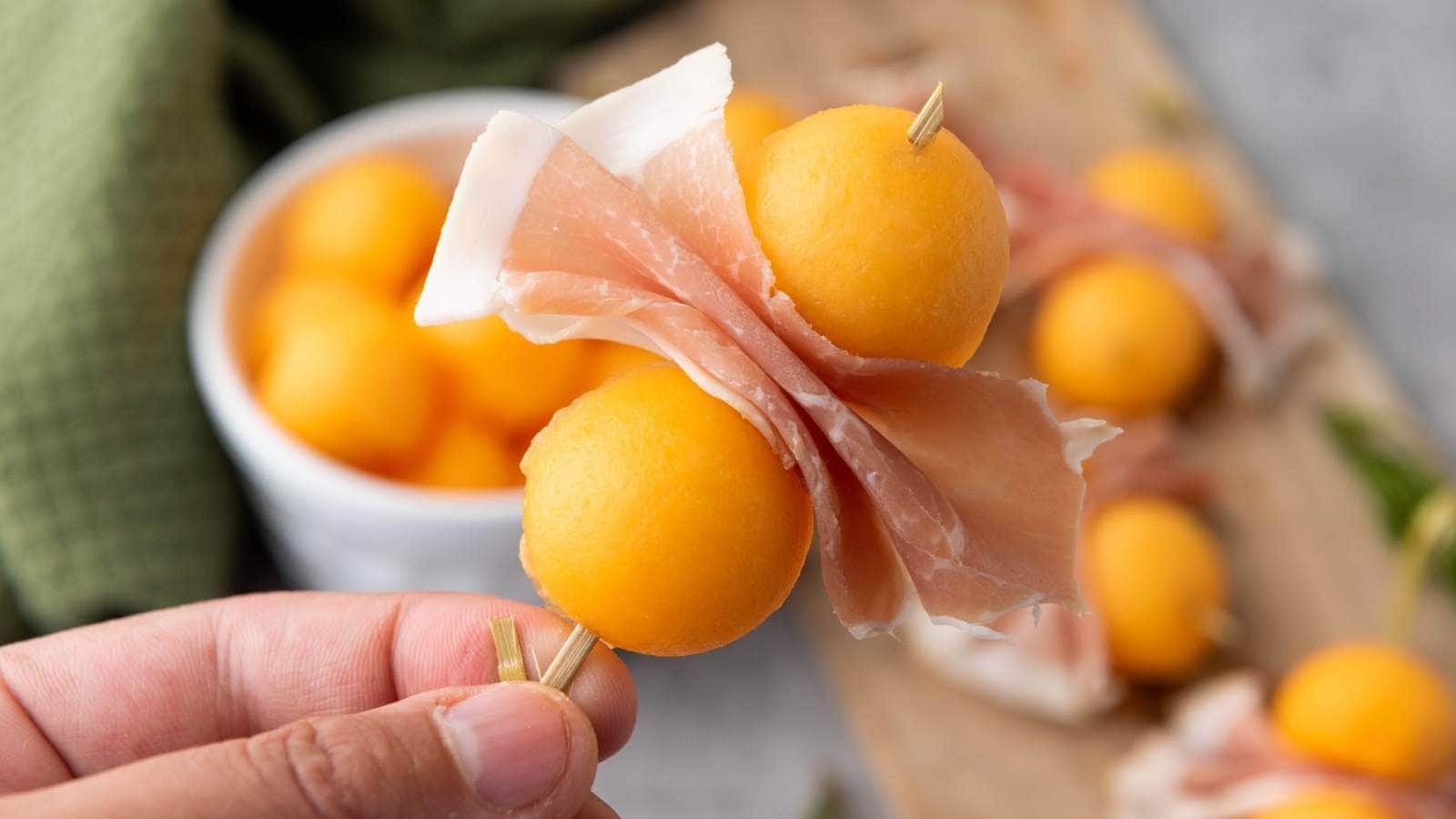 A hand holds a skewer with two cantaloupe balls and a folded slice of prosciutto.