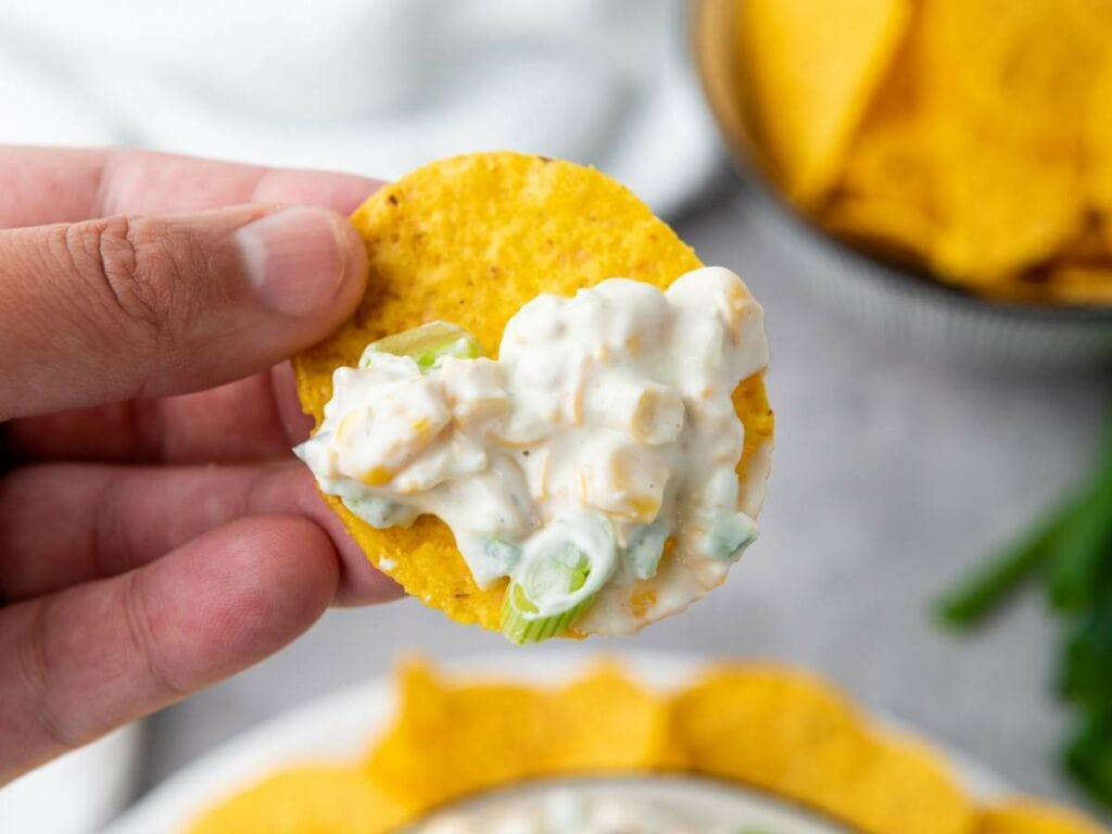 A hand holds a tortilla chip topped with a creamy corn dip.