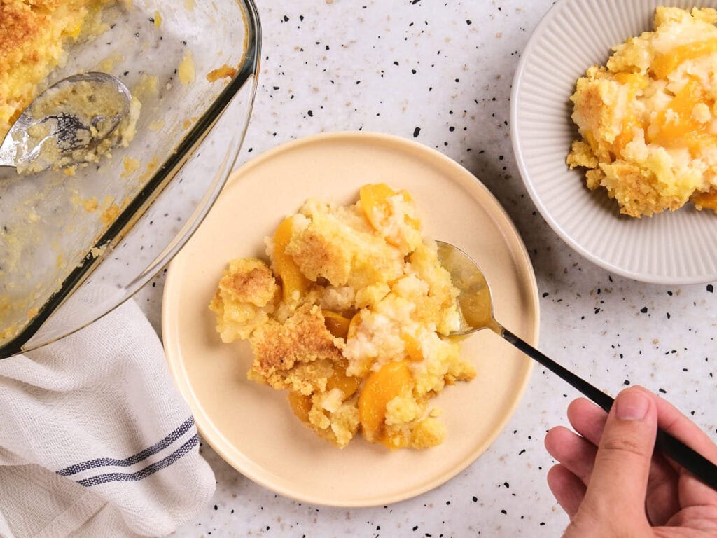 A plate with a serving of peach cobbler is shown, with a spoon and a hand holding it.