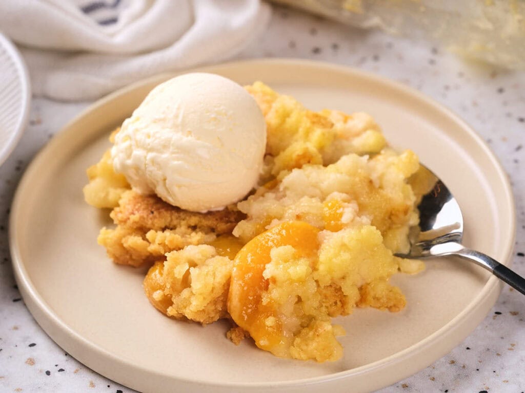 A serving of peach cobbler topped with a scoop of vanilla ice cream on a beige plate with a spoon.