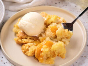 A plate with peach cobbler made with cake mix, topped with a scoop of vanilla ice cream, with a spoon.