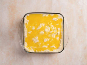 A glass baking dish filled with an unbaked mixture of butter and dry ingredients, sits on a light-colored surface.