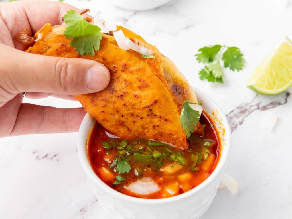 A hand dips a cheesy quesabirria tacos into a bowl of red consommé.