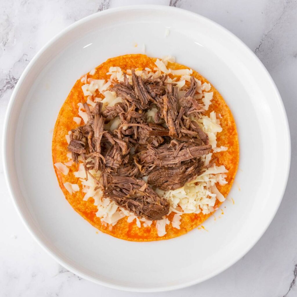 A plate with a corn tortilla topped with shredded beef and grated cheese.
