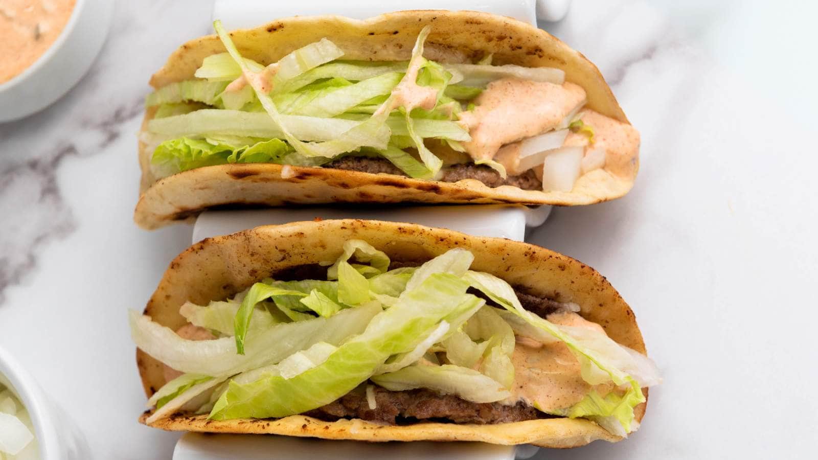 Two smash burger tacos filled with ground beef, shredded lettuce, diced onions, and a creamy sauce, served in crispy taco shells.