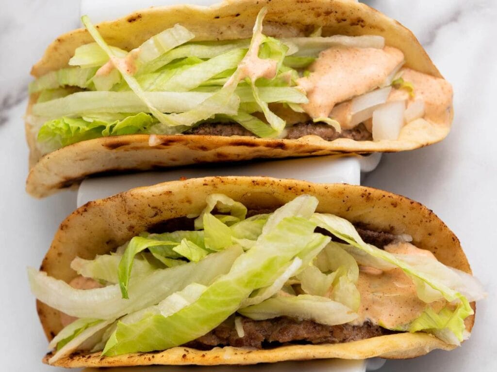 Two smash burger tacos filled with ground beef, shredded lettuce, diced onions, and a creamy sauce.