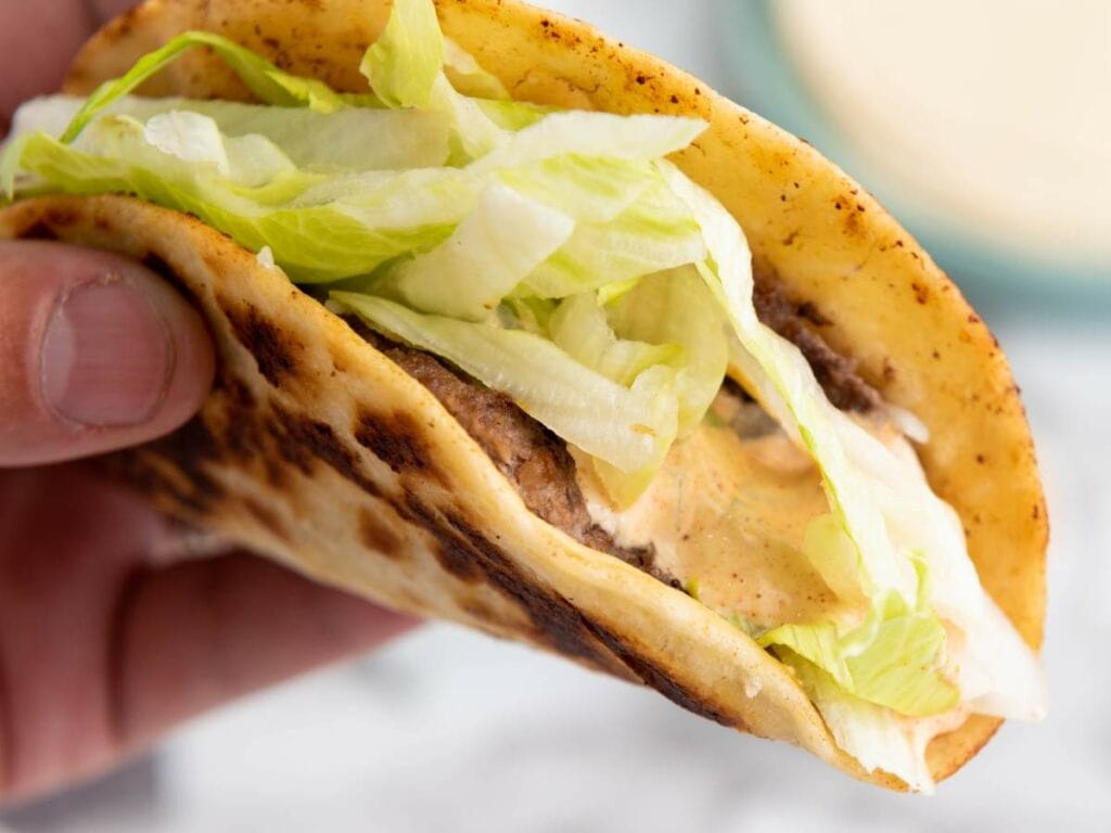 A smash burger taco with a toasted shell, filled with ground beef, shredded lettuce, and sauce.