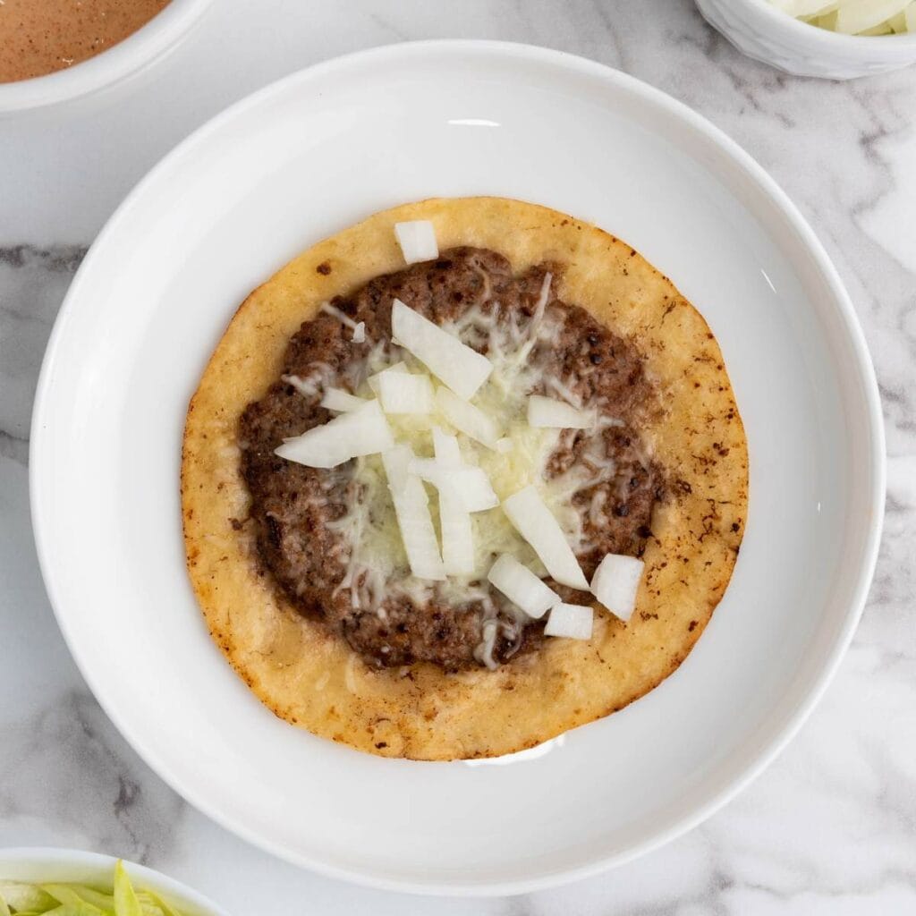 A flat, round fried tortilla topped with ground beef, melted cheese, and chopped raw onions.