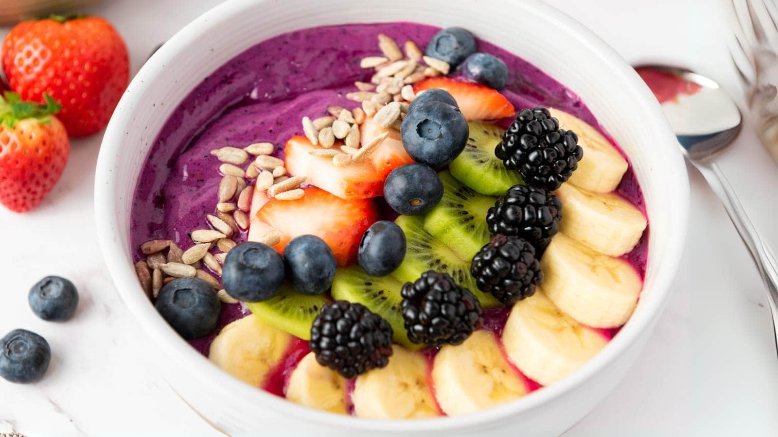 A smoothie bowl topped with sliced banana, kiwi, strawberries, blackberries, blueberries, and sunflower seeds.