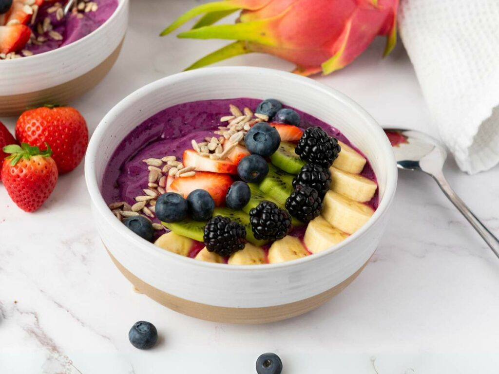 A bowl of purple smoothie topped with banana slices, blueberries, blackberries, kiwi, strawberries, and sunflower seeds.