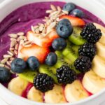 A smoothie bowl topped with sliced bananas, kiwi, strawberries, blackberries, blueberries, and sunflower seeds.