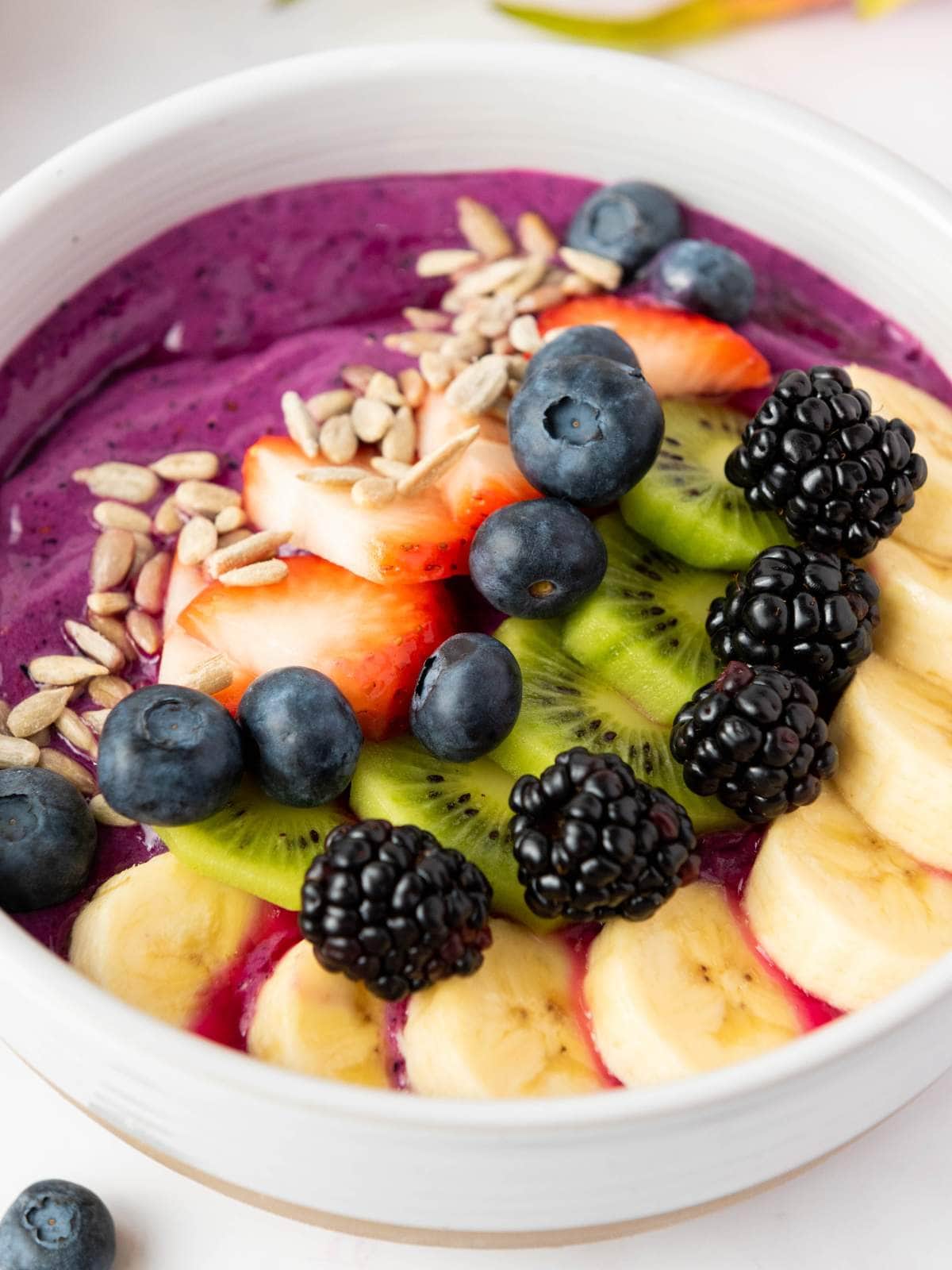 A smoothie bowl topped with sliced bananas, kiwi, strawberries, blackberries, blueberries, and sunflower seeds.