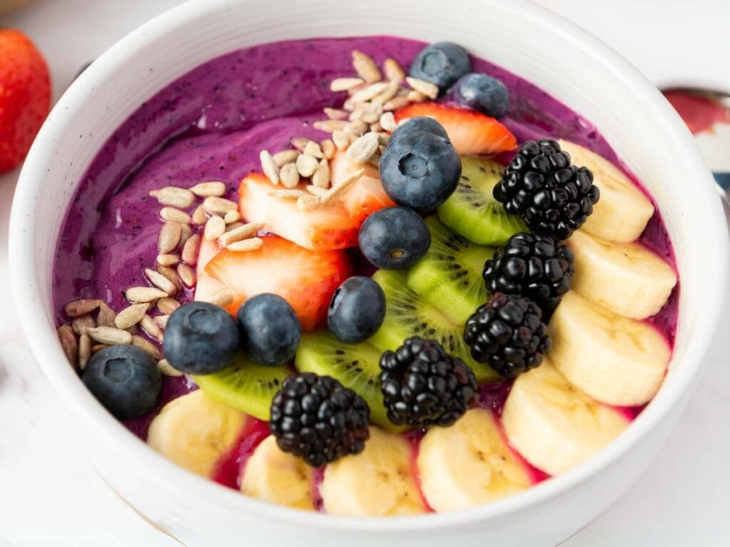 A bowl of purple smoothie topped with banana slices, kiwi, blackberries, blueberries, strawberries, and sunflower seeds.
