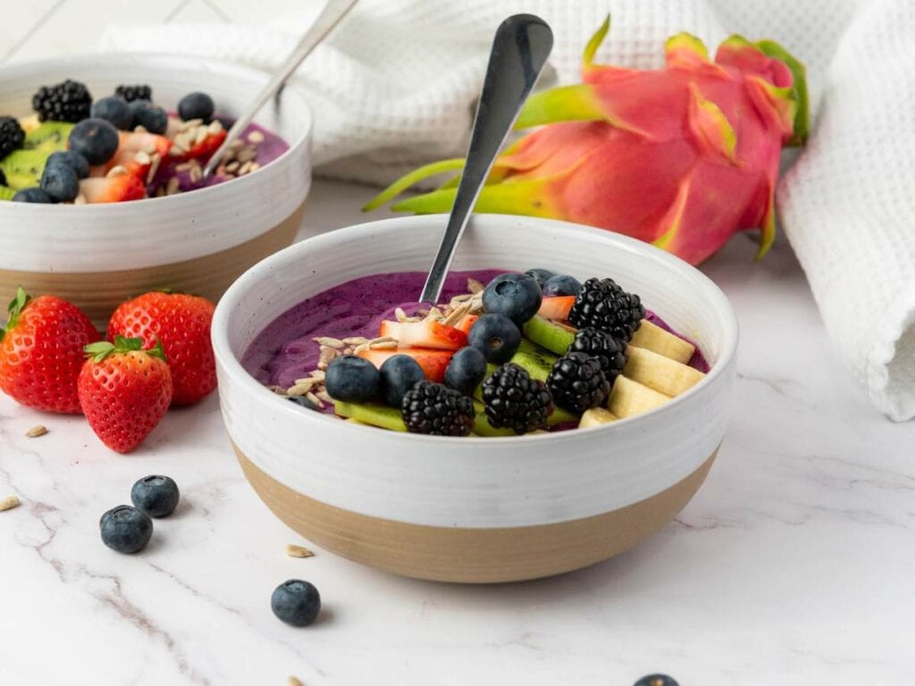 A bowl of purple smoothie topped with sliced banana, kiwi, blackberries, blueberries, and coconut.
