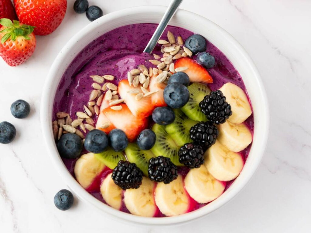 A smoothie bowl topped with banana slices, kiwi, blackberries, blueberries, strawberries, and sunflower seeds.