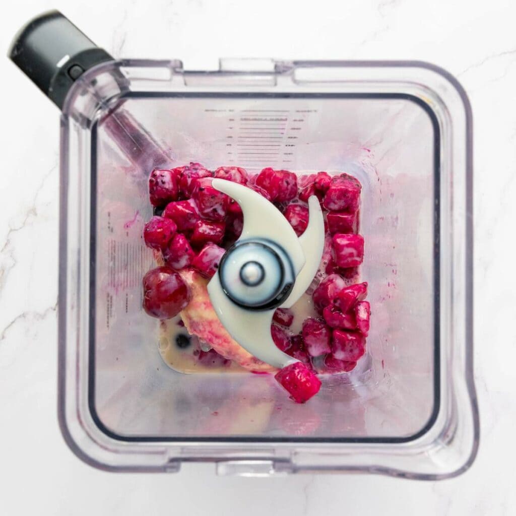 Overhead view of a blender containing frozen cherries, banana, and milk, ready to be blended.
