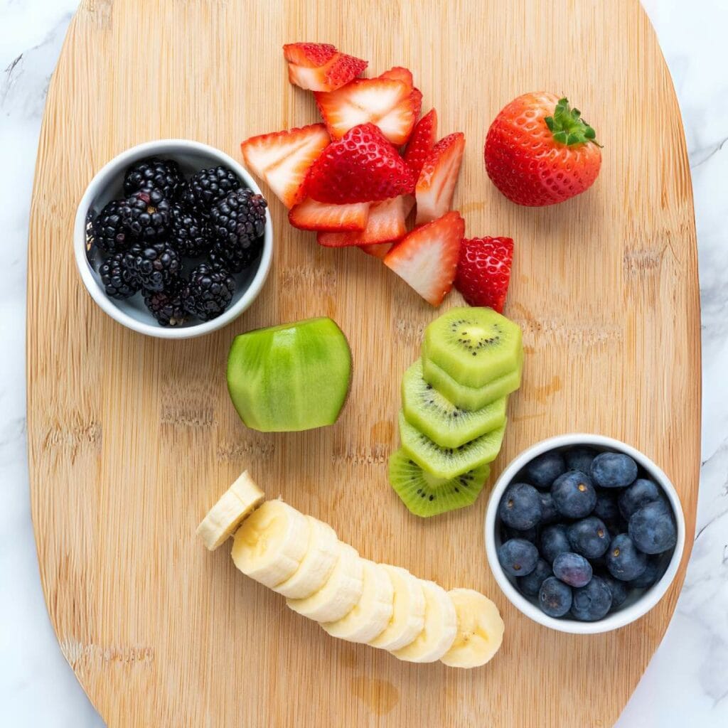 A wooden board with sliced strawberries, kiwis, and bananas, a whole strawberry, and small bowls of blackberries and blueberries.