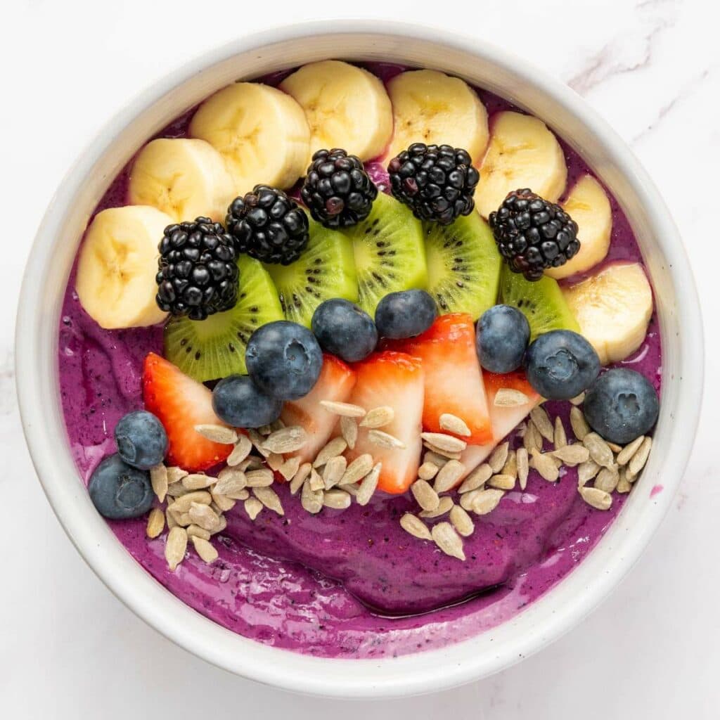 A smoothie bowl topped with banana slices, kiwi, blackberries, blueberries, strawberries, and sunflower seeds, served in a white bowl on a white surface.