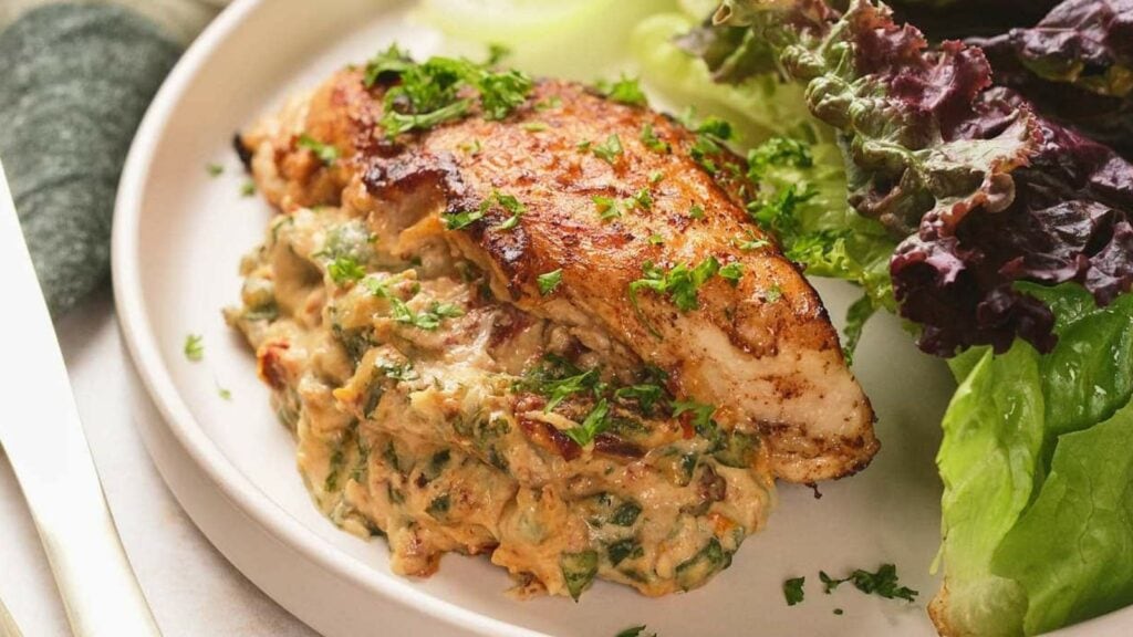 A plate with seasoned grilled chicken breast on top of a creamy spinach mixture, served with leafy greens on the side.