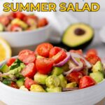 A bowl of avocado and tomato salad with red onion and cucumber, set on a counter with a halved avocado and lemon in the background.