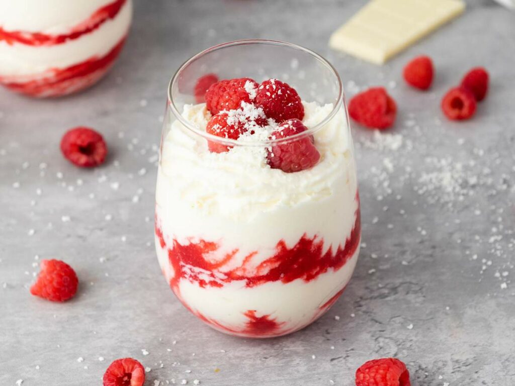A glass of white chocolate mousse and red raspberry puree, topped with raspberries and white chocolate shavings.