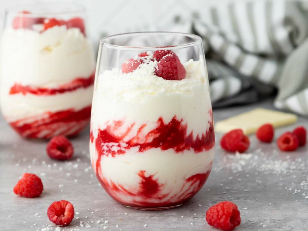 A glass of white chocolate mousse filled with layers of whipped cream and raspberry sauce, topped with fresh raspberries and white chocolate shavings