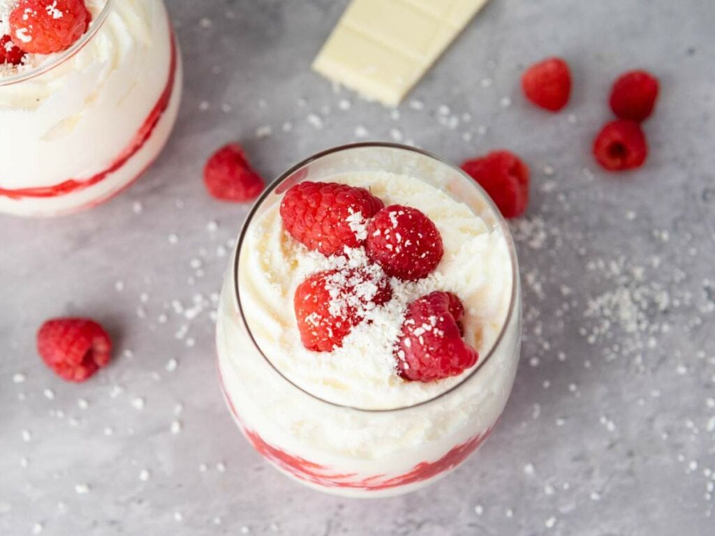 A glass of white chocolate mousse filled with creamy dessert topped with raspberries and white chocolate shavings.